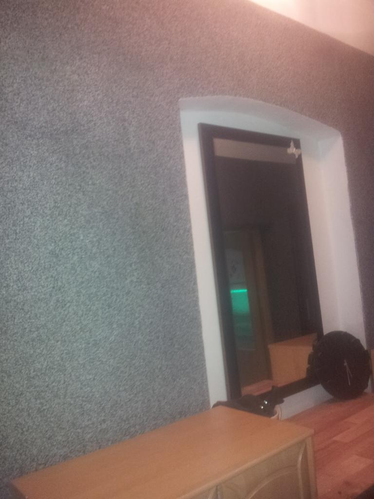 Szara ściana z dekoracyjną strukturą, wnęka z lustrem w ciemnej ramie i białą obudową, widoczny fragment drewnianej podłogi i mebla.