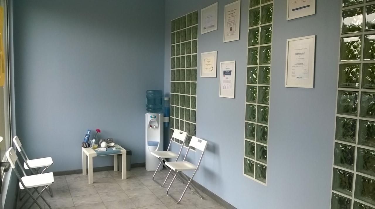 Jasne pomieszczenie biurowe z błękitnymi ścianami, dekoracyjnymi panelami z luksferów, białymi krzesłami, stolikiem z kwiatami i dyplomami na ścianie.