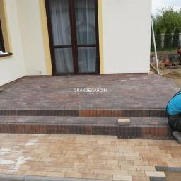 Grande Gardini Maciej Harendarczyk - Etap prac: układanie wielokolorowej kostki brukowej na schodach wejściowych do domu, widoczni dwaj pracownicy w trakcie pracy oraz ubijarka w tle.