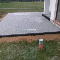 Szary taras z kostki brukowej ułożony przy domu, z czarnym obrzeżem i wystającą rurą kanalizacyjną na trawniku.