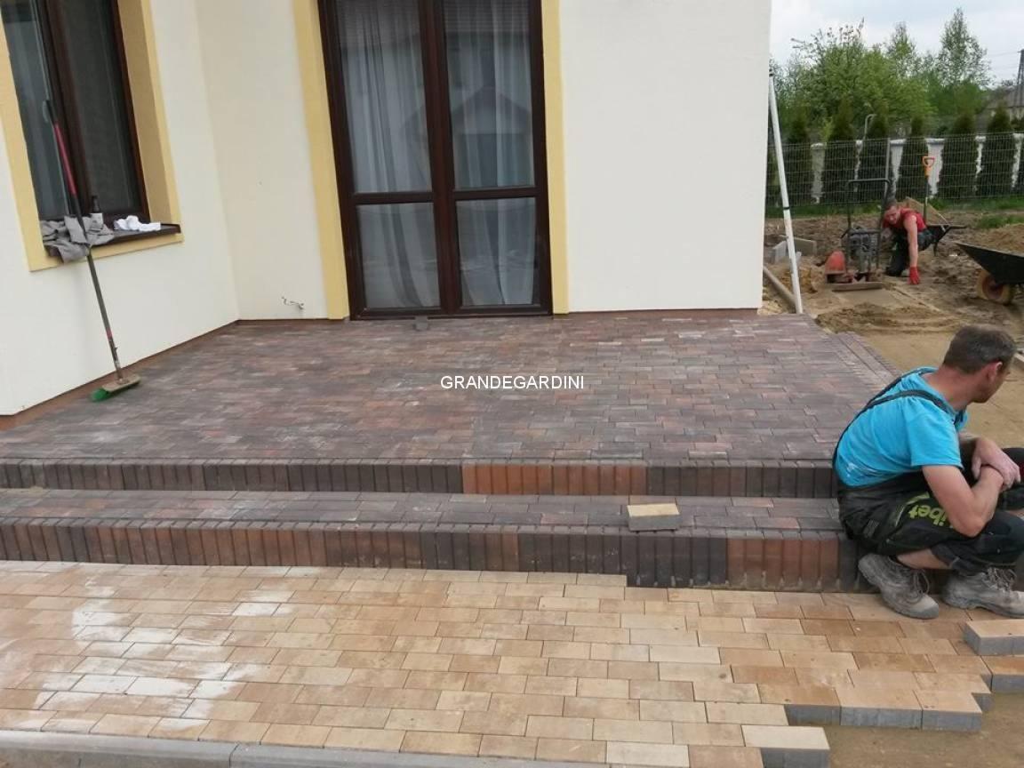 Etap prac: układanie wielokolorowej kostki brukowej na schodach wejściowych do domu, widoczni dwaj pracownicy w trakcie pracy oraz ubijarka w tle.