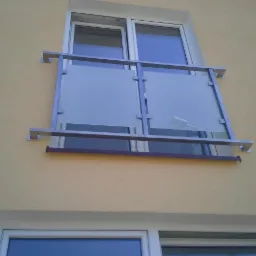 Balkon z nowoczesną balustradą ze stali i szkła matowego, zamontowaną na elewacji budynku w kolorze piaskowym, widok z dołu.