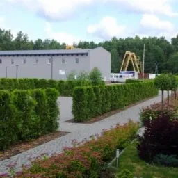 Widok na zakład prefabrykacji betonowej z placem składowym, zielenią ozdobną i drogą dojazdową wyłożoną kostką brukową.