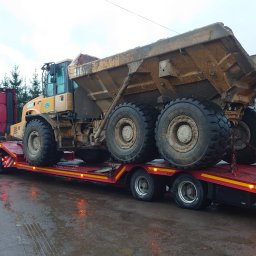 ZoomZoom Service Albert Magier - Czerwony ciągnik siodłowy z naczepą typu platforma przewozi żółtą, brudną wywrotkę budowlaną na tle szarego nieba i drzew bez liści.