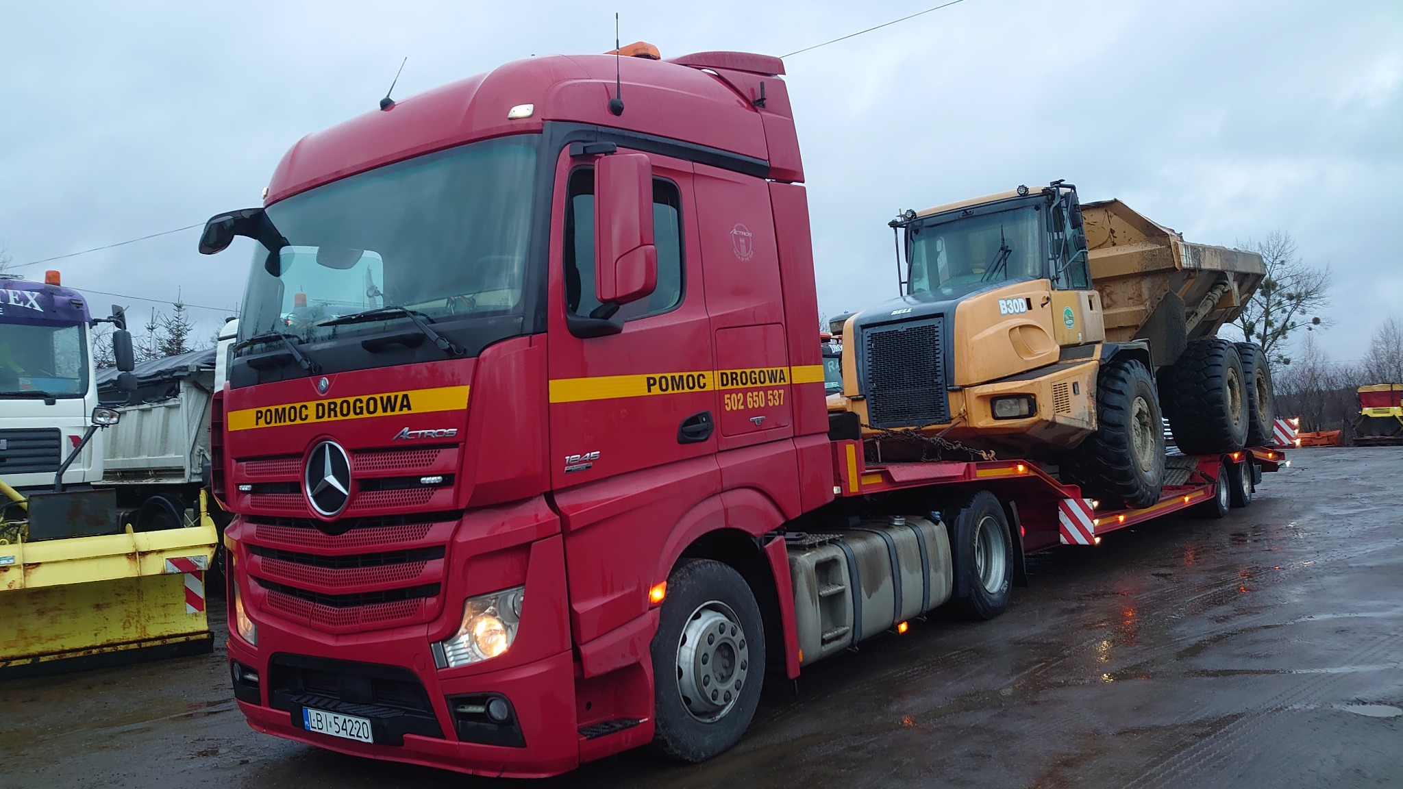 Czerwona ciężarówka pomocy drogowej Mercedes-Benz Actros z żółtym wozidłem budowlanym Bell B30D na niskopodwoziowej naczepie, zaparkowana na mokrym placu.