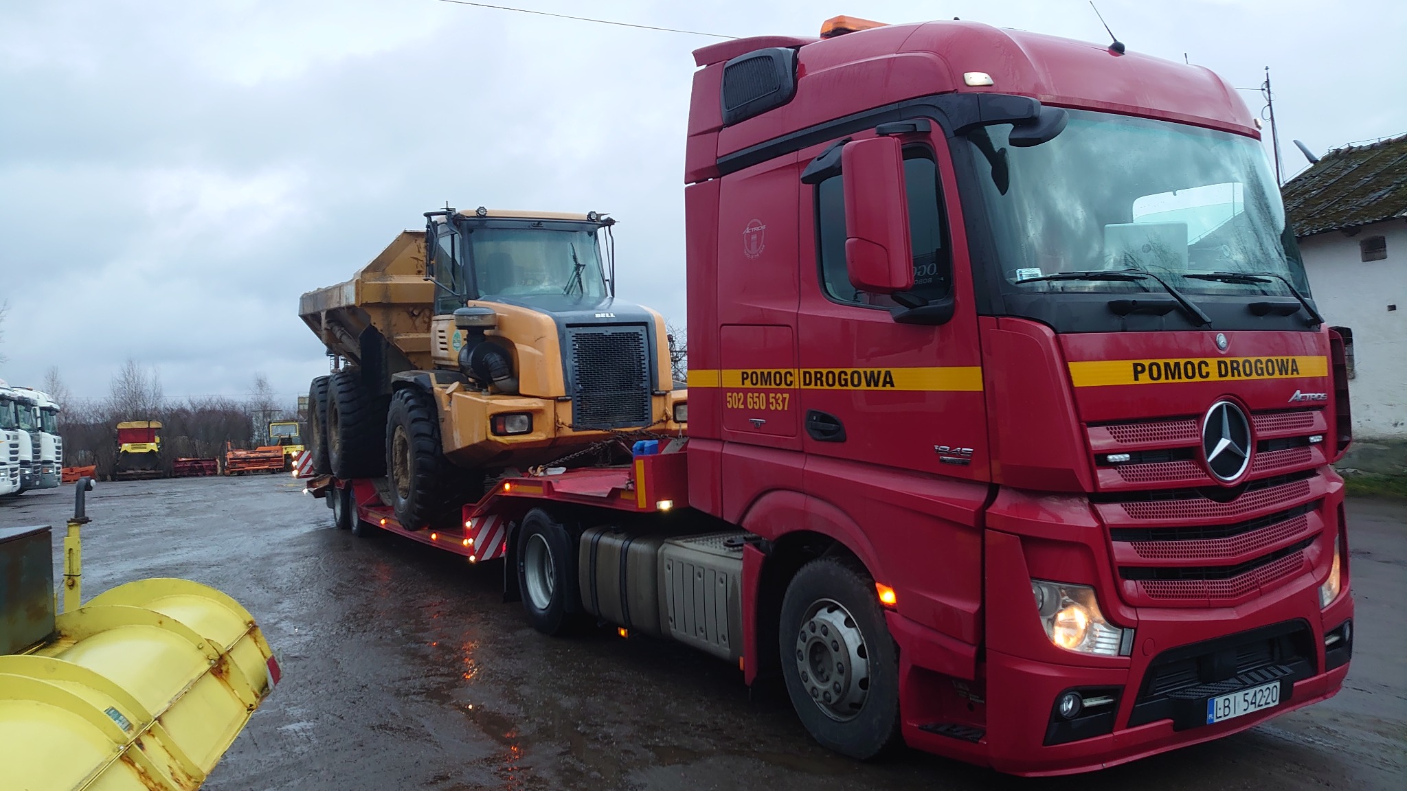 Czerwony ciągnik siodłowy Mercedes-Benz z napisem 'POMOC DROGOWA' przewozi na lawecie żółty wozidło przegubowe Bell pod pochmurnym niebem.