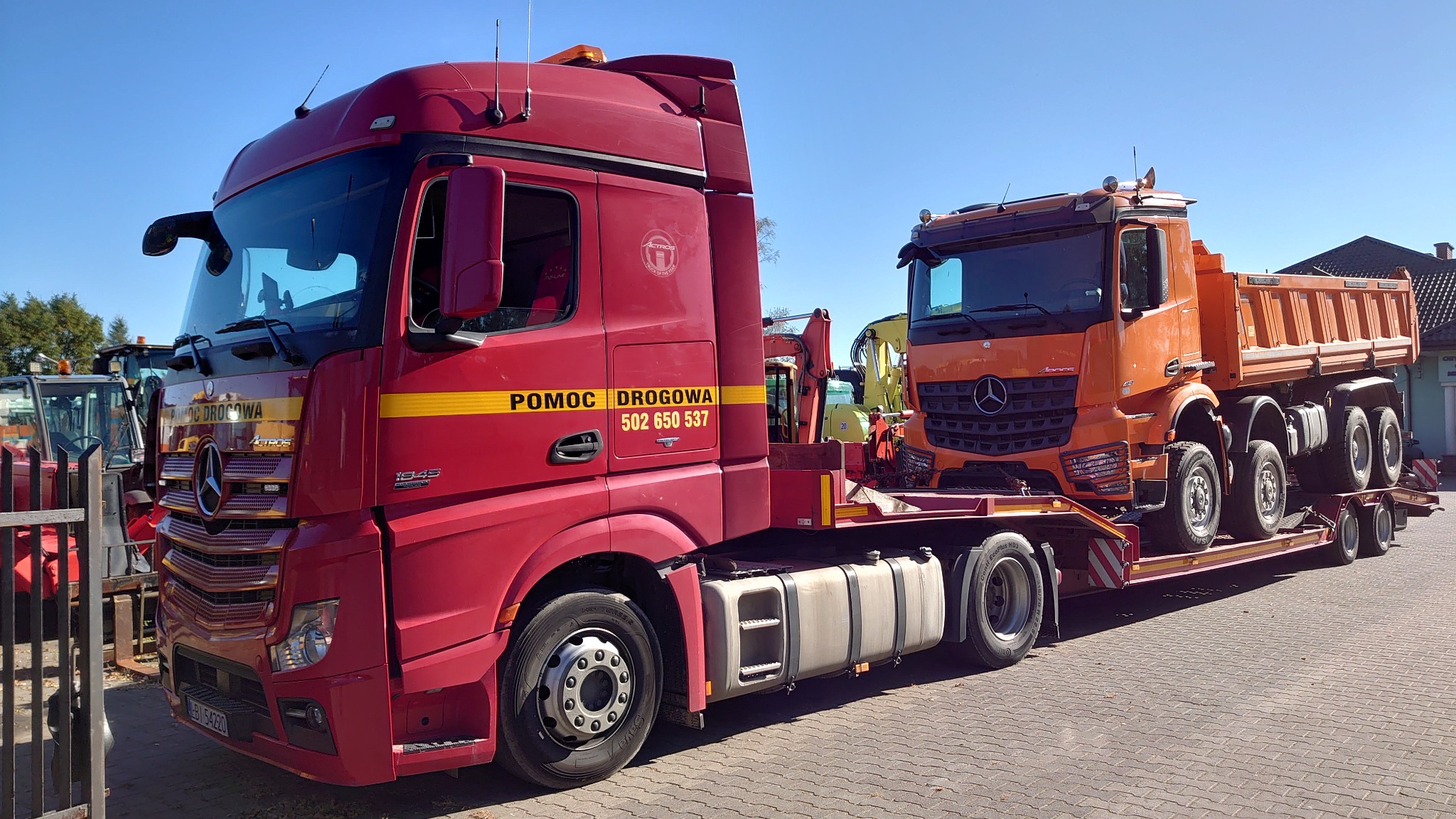 Czerwona laweta Mercedes-Benz z napisem 'Pomoc Drogowa' przewozi pomarańczową wywrotkę Mercedes-Benz Actros na tle kostki brukowej i błękitnego nieba.