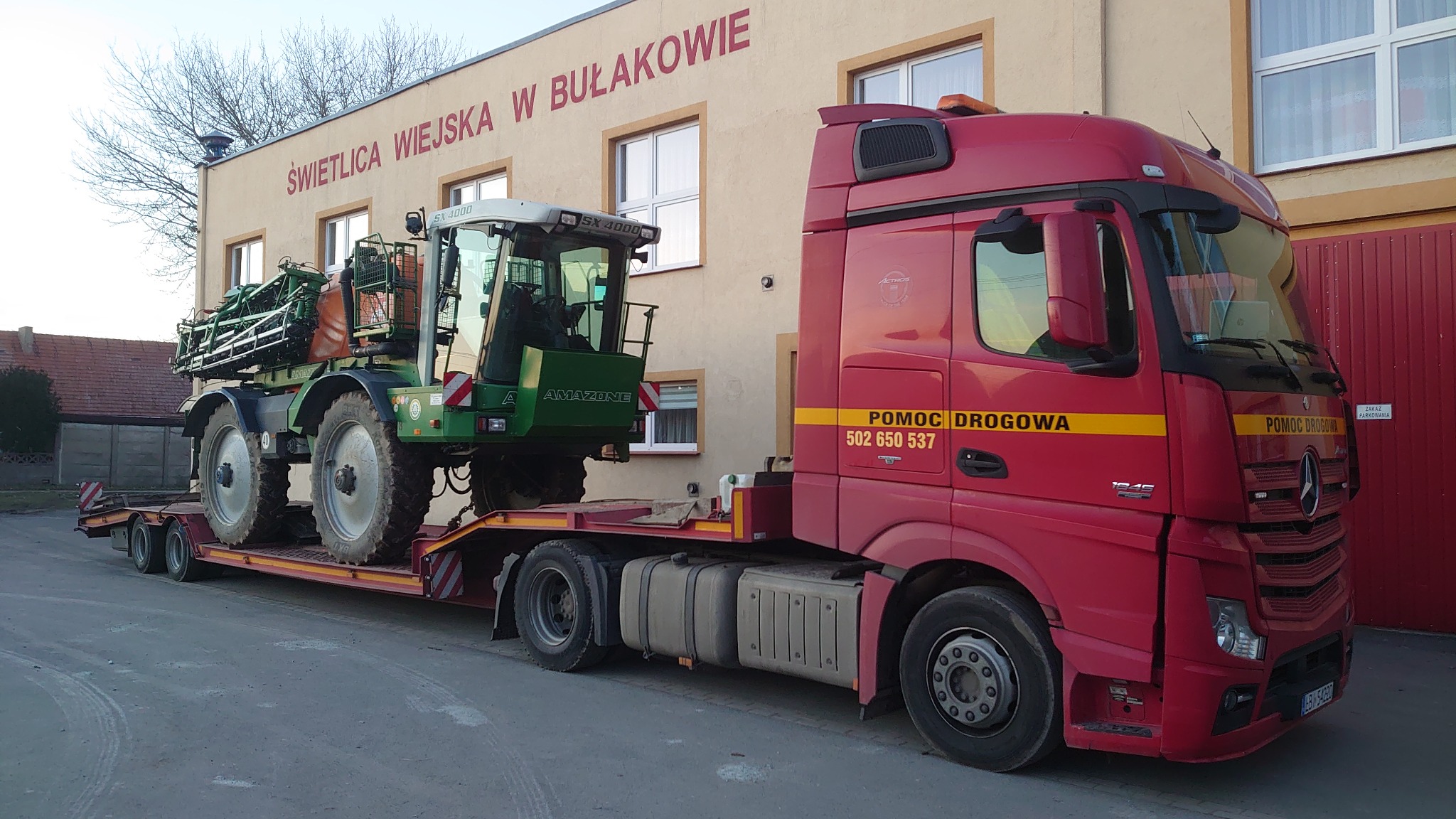 Czerwona ciężarówka z naczepą niskopodwoziową przewożąca zielony opryskiwacz polowy marki Amazone SX 4000 na tle budynku 'Świetlica Wiejska w Bułakowie'.