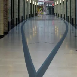 Długi korytarz z błyszczącą podłogą wyłożoną płytkami w odcieniach szarości, z ciemnoszarym wzorem w kształcie strzały biegnącym wzdłuż środka, oświetlony rzędami lamp ściennych.