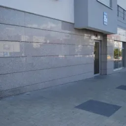 Elewacja budynku wyłożona szarymi płytami kamiennymi o drobnej fakturze, z widocznym wejściem i chodnikiem z kostki brukowej.