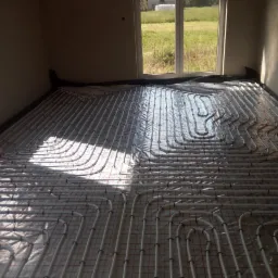 Rozłożona instalacja ogrzewania podłogowego z białych rurek na folii z czerwoną siatką, widoczne okno i fragment schodów.