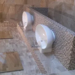 Dwie nowoczesne, białe umywalki wkomponowane w mozaikową ściankę, z elementami płytek imitujących marmur na podłodze i ścianach, wykończone w odcieniach beżu i brązu.