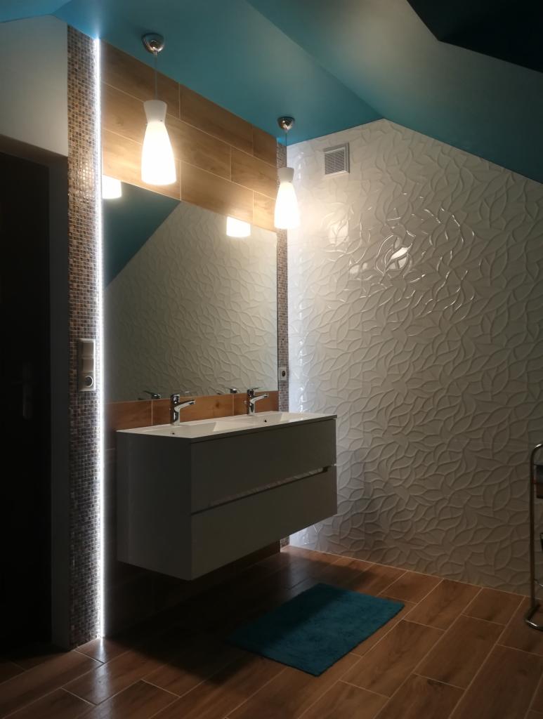 Nowoczesna łazienka z podwójną umywalką, oświetleniem LED wzdłuż ściany z mozaiki, białą ścianą z dekoracyjnym wzorem liści, drewnopodobną podłogą i turkusowym akcentem w postaci małego dywanika.