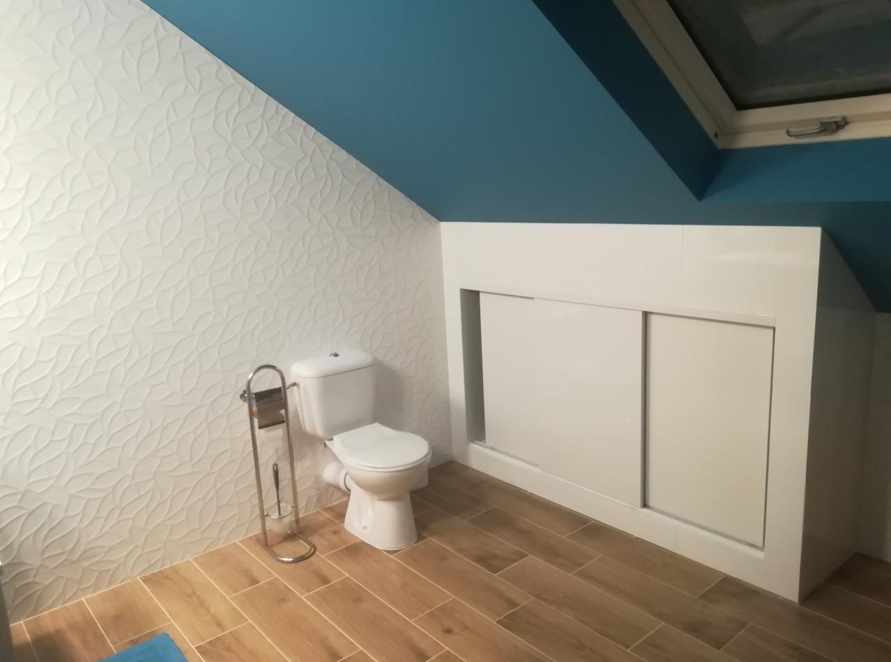 Wykończona łazienka na poddaszu ze skośnym sufitem pomalowanym na niebiesko, białą toaletą, dekoracyjnymi białymi płytkami na ścianie i szafką w zabudowie z przesuwnymi drzwiami.