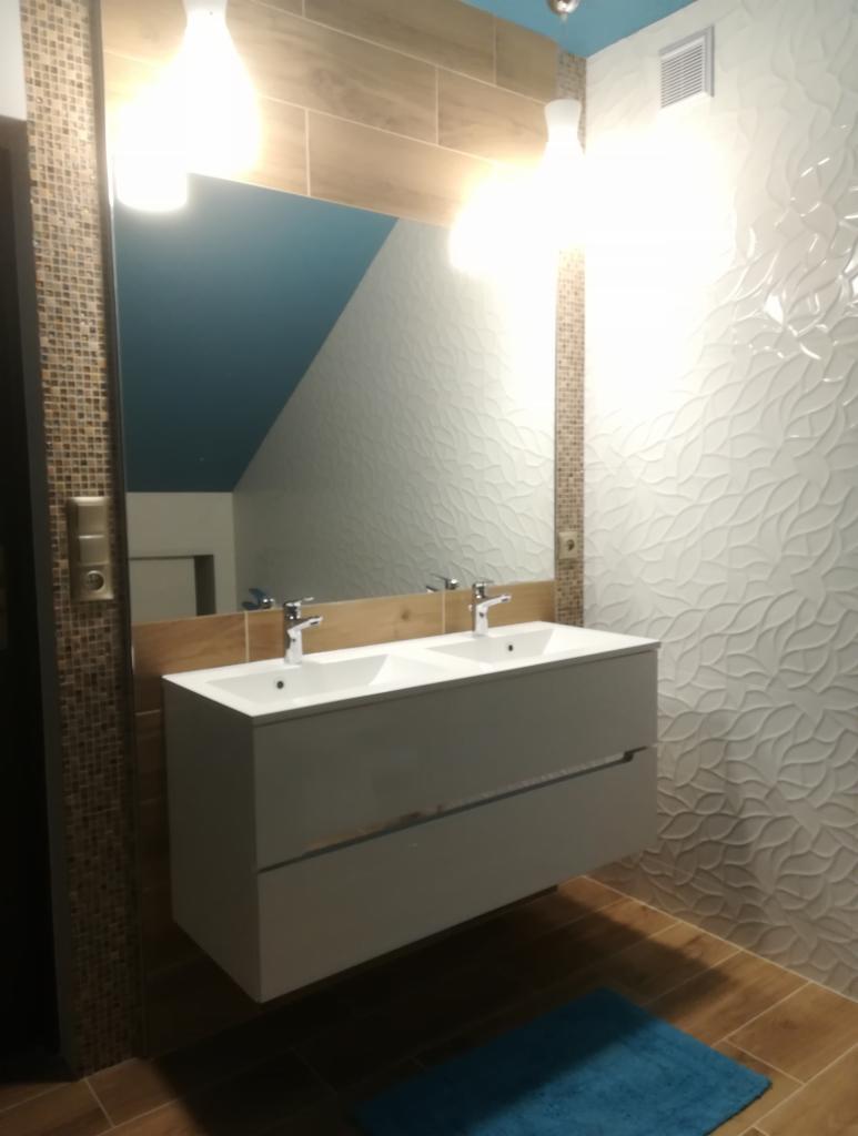Nowoczesna łazienka z podwójną białą umywalką na szafce, wzorzyste białe płytki na ścianie i drewnopodobne płytki na podłodze, odbicie błękitnego sufitu w lustrze.