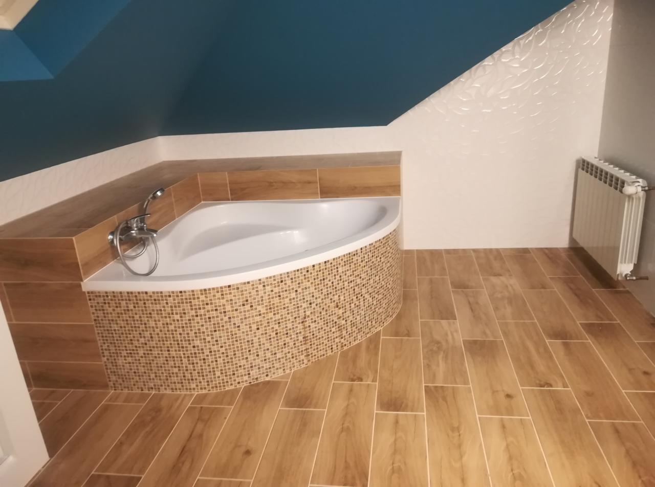 Wykończona łazienka na poddaszu z narożną wanną obudowaną mozaiką, drewnopodobne płytki na podłodze i ścianie, biała ściana z dekoracyjnym wzorem i ciemnoniebieski sufit.