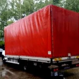 Czerwony bus z plandeką zaparkowany na mokrym placu, widok z tyłu, zarys budynków i drzew w tle.