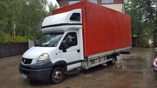 Biały samochód dostawczy Renault z czerwoną plandeką na mokrym brukowanym podjeździe przed domem.