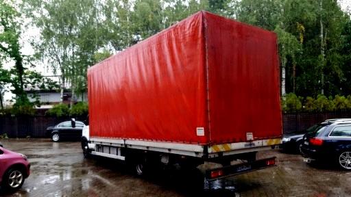 Czerwony bus z plandeką zaparkowany na mokrym placu, widok z tyłu, zarys budynków i drzew w tle.