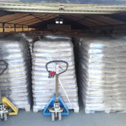 Załadowany magazyn pelletu w workach na paletach, zabezpieczonych folią, z wózkami paletowymi w kolorach żółtym i niebieskim z czerwonymi rękawicami roboczymi na uchwytach.