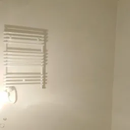 Biały, drabinkowy grzejnik łazienkowy zamontowany na świeżo pomalowanej, kremowej ścianie w narożniku łazienki, z widocznymi śladami po niedawnym malowaniu.