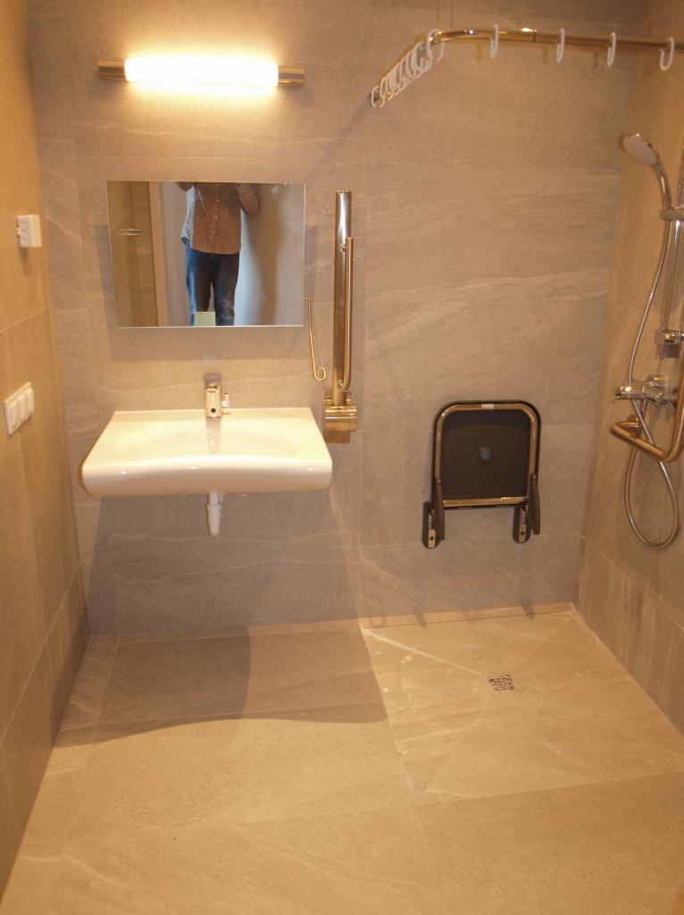 Jasna łazienka z beżowymi płytkami, umywalką wiszącą, składanym krzesełkiem prysznicowym i relingiem na ręczniki, z widocznym odbiciem osoby w lustrze.