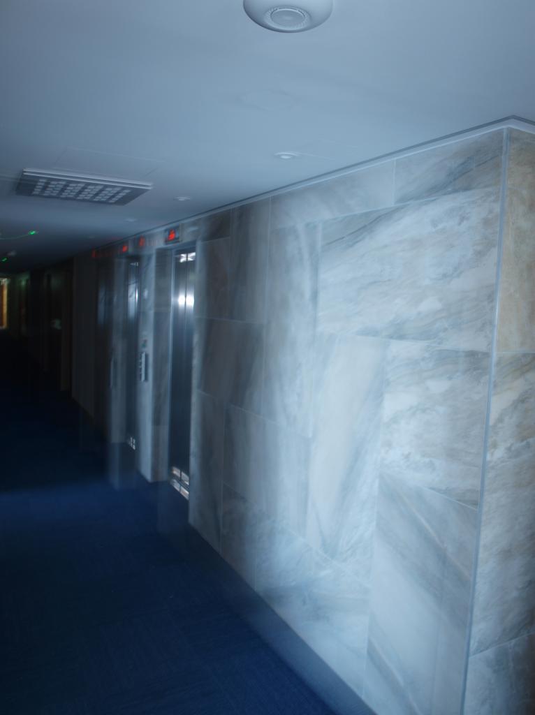 Korytarz w budynku z wykończonymi ścianami w marmurze, widoczne trzy windy z numerami pięter.