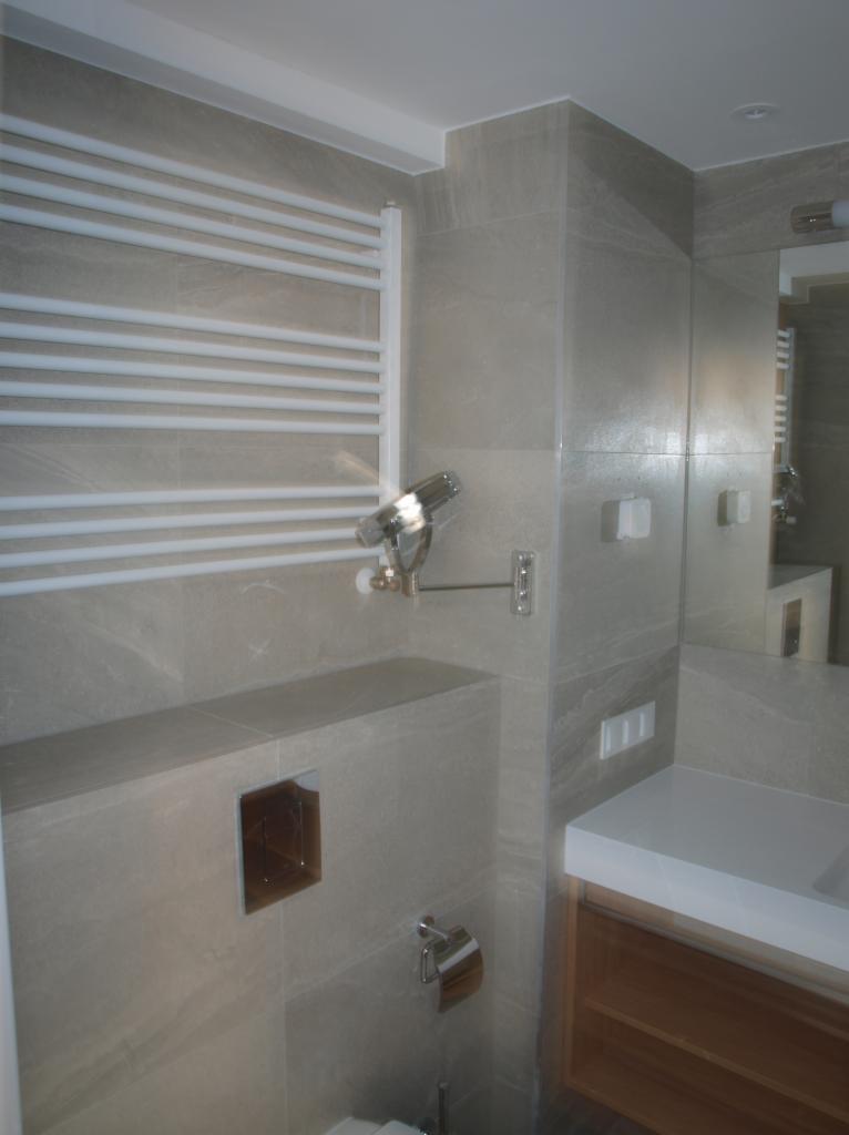 Wykończona łazienka z szarymi płytkami imitującymi kamień, białym grzejnikiem drabinkowym, chromowaną armaturą i zabudowaną toaletą z wnęką na akcesoria.