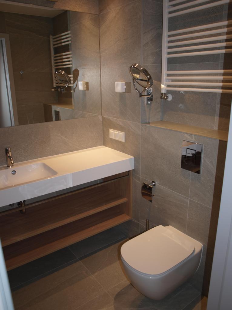 Nowoczesna łazienka z szarymi płytkami imitującymi kamień, wisząca toaleta, umywalka nablatowa z szafką z otwartymi półkami, grzejnik drabinkowy i lustro powiększające.