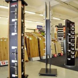 Ekspozycja klamek do drzwi w sklepie budowlanym, widoczne stojaki z różnymi modelami klamek na tle desek podłogowych.