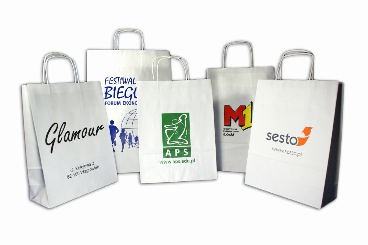 Białe papierowe torby z nadrukami reklamowymi różnych firm i wydarzeń, ustawione na białym tle, prezentujące różnorodność wzorów i kolorów.