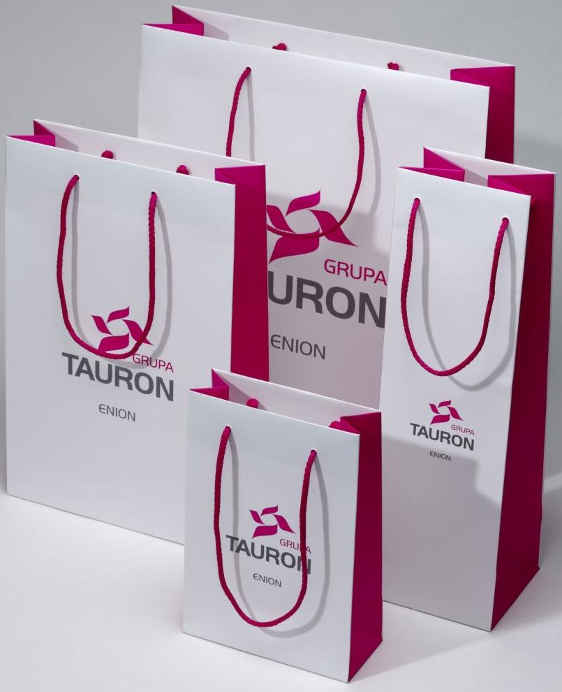 Białe torby papierowe z różowymi uchwytami i nadrukowanym logo firmy Tauron Enion, różne rozmiary, ustawione na jasnym tle.