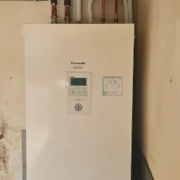 Biała pompa ciepła Panasonic Aquarea z podłączonymi rurami miedzianymi i izolowanymi, zamontowana na tle surowej ściany w trakcie remontu.