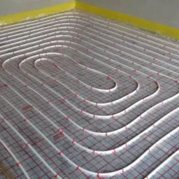 Białe rury ogrzewania podłogowego ułożone w regularne pętle na szarej, zabezpieczonej folią podłodze, widoczne czerwone elementy mocujące.