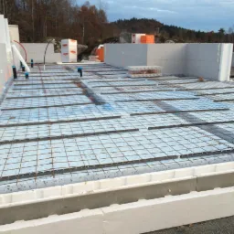 Budowa fundamentów domu z wykorzystaniem szalunków styropianowych i zbrojenia stalowego, widoczne instalacje pionowe i zapasy materiałów budowlanych w tle.