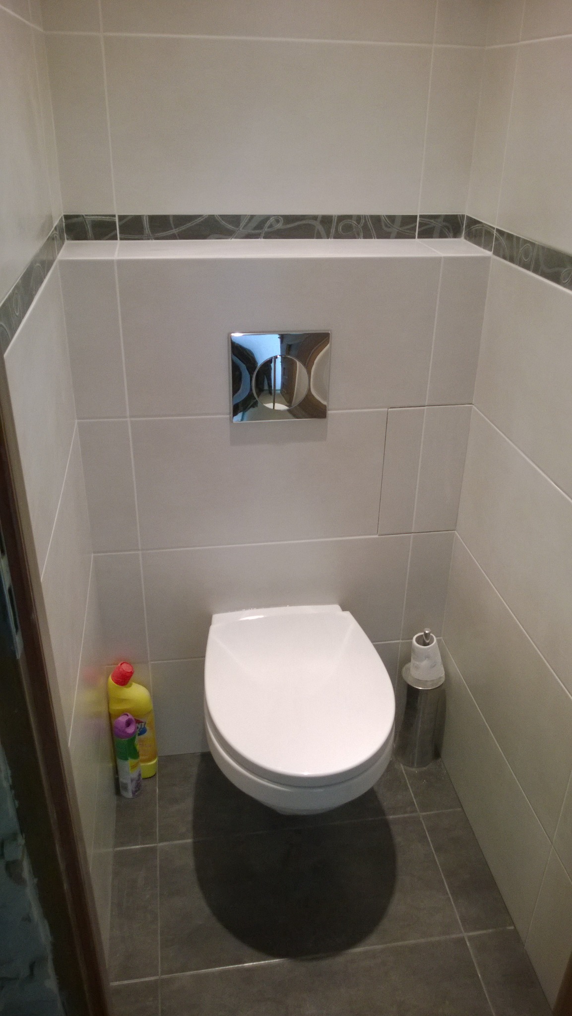 Nowoczesna toaleta podwieszana w wykończonej łazience z szarymi płytkami podłogowymi i beżowymi płytkami ściennymi z dekoracyjnym pasem, widoczny chromowany przycisk spłuczki i pojemnik na papier...