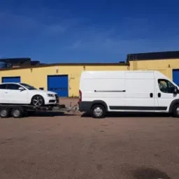 Biały samochód osobowy marki Audi A5 na przyczepie transportowej, ciągniętej przez biały samochód dostawczy na tle żółtego budynku z niebieskimi bramami garażowymi w słoneczny dzień.