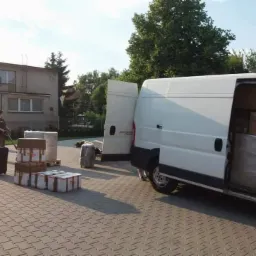 Pracownik na zewnątrz białego busa z otwartymi drzwiami, ładuje kartony i walizki na palecie na chodniku z kostki brukowej przed budynkiem.