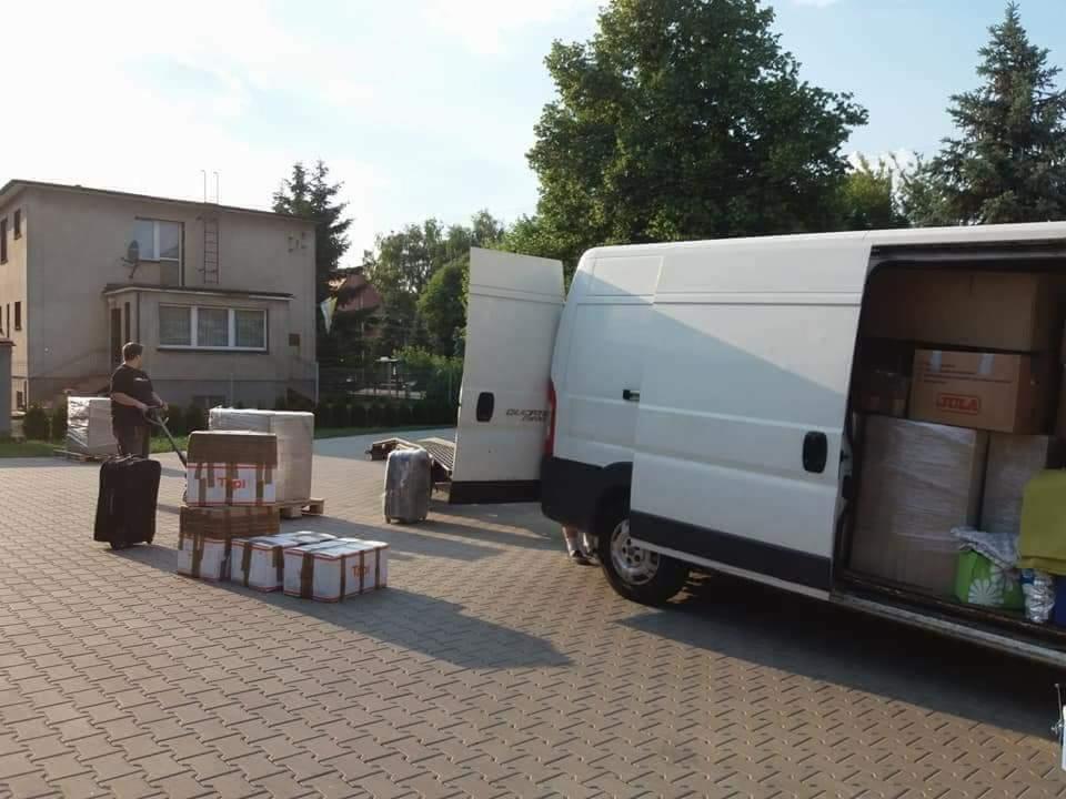 Pracownik na zewnątrz białego busa z otwartymi drzwiami, ładuje kartony i walizki na palecie na chodniku z kostki brukowej przed budynkiem.