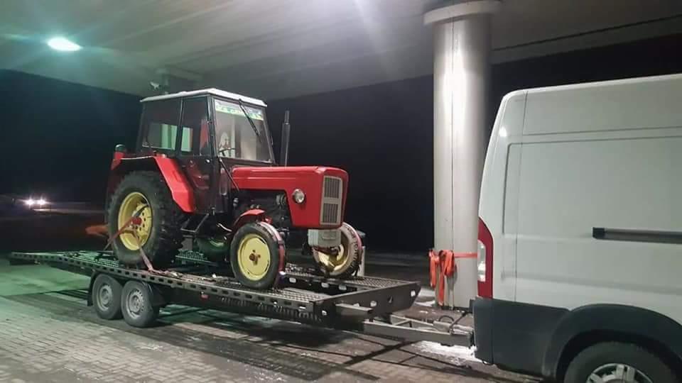 Czerwony traktor Ursus na niskopodwoziowej przyczepie, zabezpieczony pasami, obok biały bus przy kolumnie stacji benzynowej.