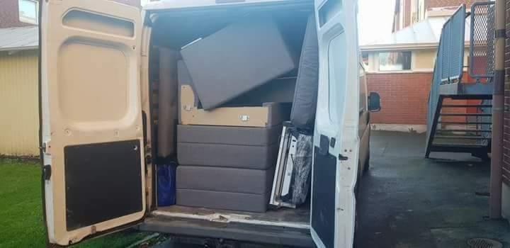 Wnętrze białego busa załadowane meblami i kartonami podczas przeprowadzki, widoczne otwarte tylne drzwi i fragment otoczenia budynku.