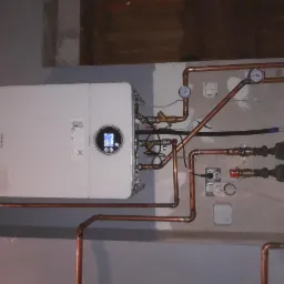 Biały piec CO marki Bosch zamontowany na szarej ścianie z widocznymi miedzianymi rurami, manometrami i elementami instalacji elektrycznej.