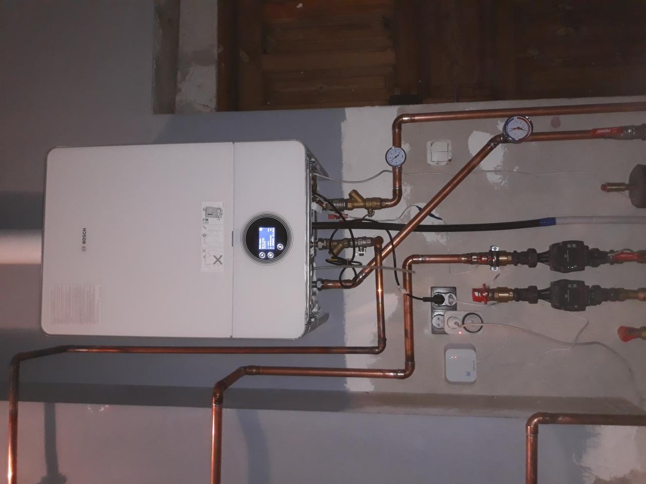 Biały piec CO marki Bosch zamontowany na szarej ścianie z widocznymi miedzianymi rurami, manometrami i elementami instalacji elektrycznej.