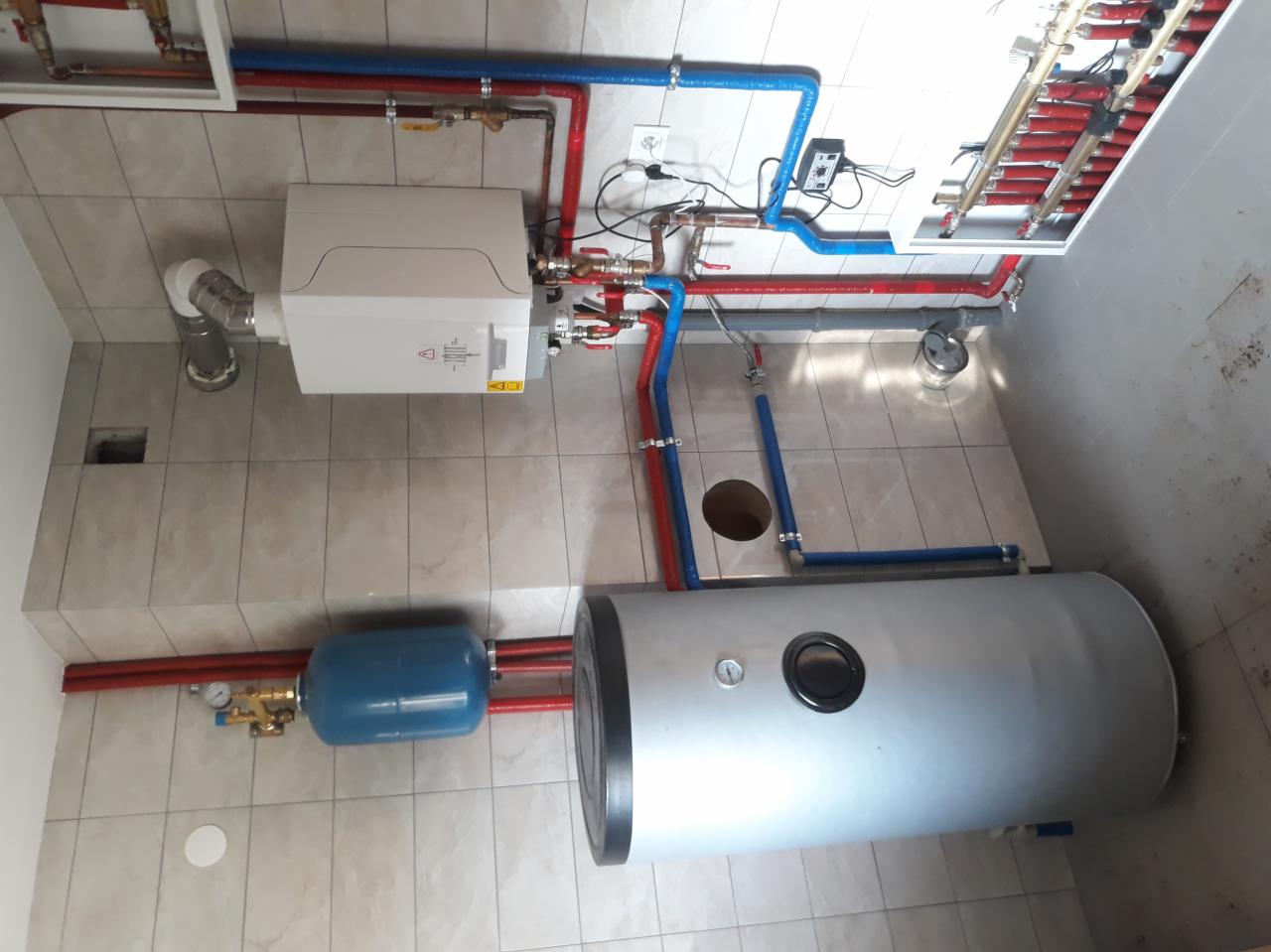 Kompletna instalacja grzewcza w kotłowni: piec, zasobnik ciepłej wody, naczynie wzbiorcze, rury w kolorach czerwonym i niebieskim, zamontowane na ścianie z płytek.
