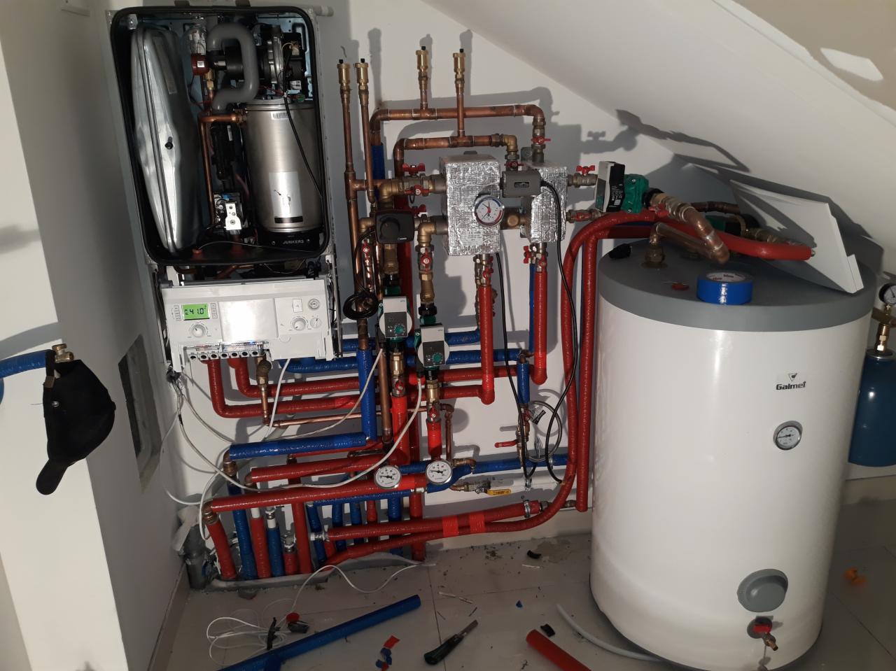 Złożona instalacja grzewcza z kotłem Junkers i zasobnikiem Galmet, widoczne liczne rury miedziane i plastikowe, manometry oraz izolacja termiczna, zamontowana w pomieszczeniu ze skośnym sufitem.