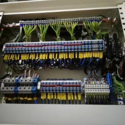 Rozdzielnica elektryczna z widocznymi przewodami oznaczonymi żółtymi etykietami, listwami zaciskowymi i elementami montażowymi w białej szafie. Przewody ułożone w uporządkowany sposób.