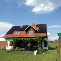 Dom jednorodzinny z panelami fotowoltaicznymi na dachu, widoczne z poziomu trawnika, z pergolą porośniętą zielenią przed tarasem.