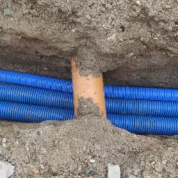 Wykop z ułożonymi niebieskimi rurami karbowanymi i pomarańczową rurą gładką, częściowo zasypane piaskiem, widoczne stopy w butach roboczych.