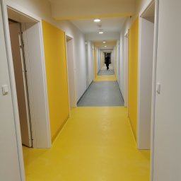 BODEN SYSTEM Wykładziny Obiektowe - Długi korytarz z jaskrawożółtą podłogą i szarą strefą, białe ściany z żółtymi panelami, białe drzwi, osoba w oddali.