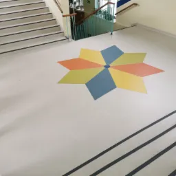 Jasna podłoga w holu z wzorem kwiatu w kolorach niebieskim, żółtym i pomarańczowym, widok z góry na schody z czarnymi paskami antypoślizgowymi.